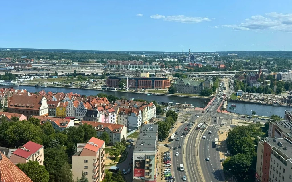 Panorama miasta z mostem nad rzeką, charakterystycznymi kolorowymi kamienicami, rozległą infrastrukturą drogową i rozbudowanym zapleczem kolejowym w tle, podkreślającym miejski krajobraz.