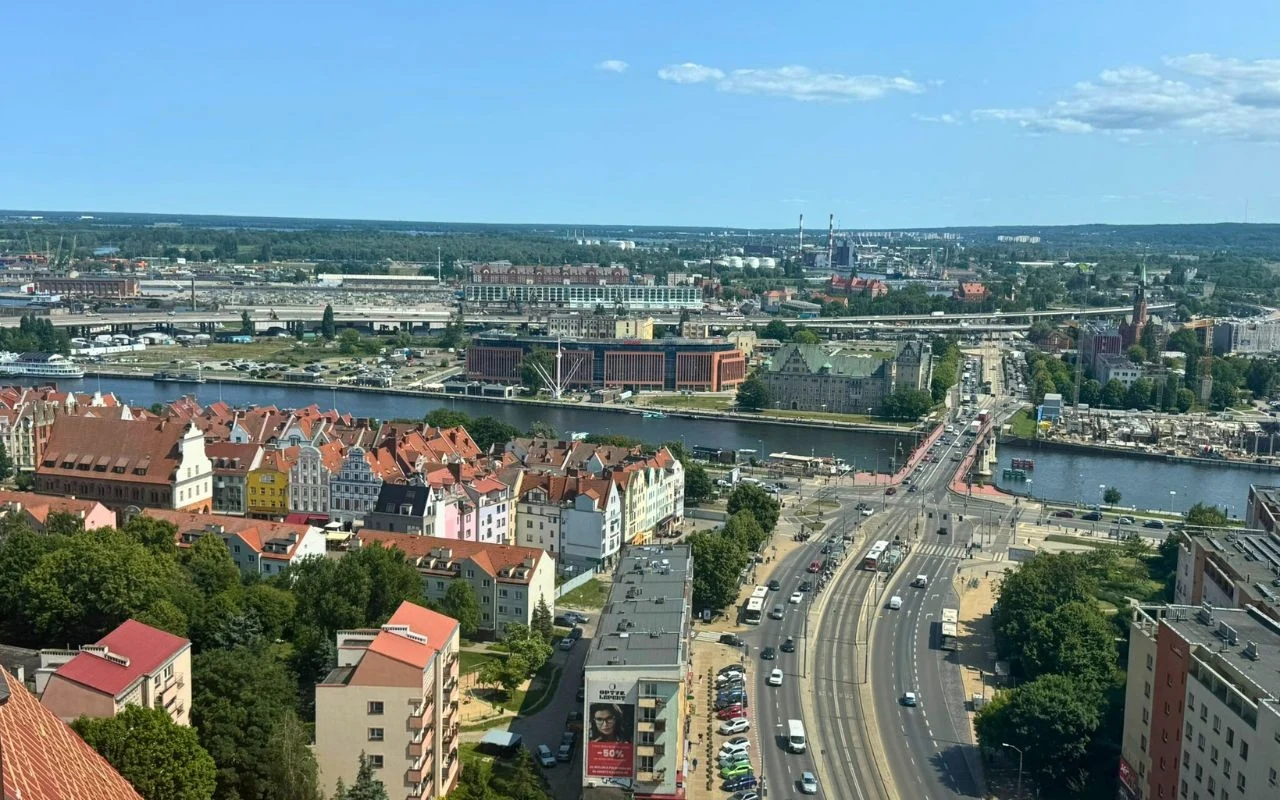 Panorama miasta z mostem nad rzeką, charakterystycznymi kolorowymi kamienicami, rozległą infrastrukturą drogową i rozbudowanym zapleczem kolejowym w tle, podkreślającym miejski krajobraz.
