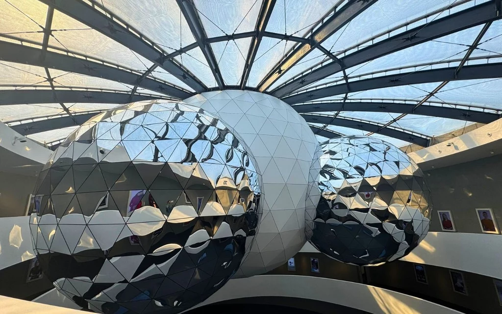 Futurystyczne, geometryczne instalacje złożone z kul o lustrzanych i trójkątnych powierzchniach w nowoczesnym wnętrzu pod dużą, przeszkloną kopułą, wokół wnętrza widoczne są biegnące balkony i oprawy eksponatów.