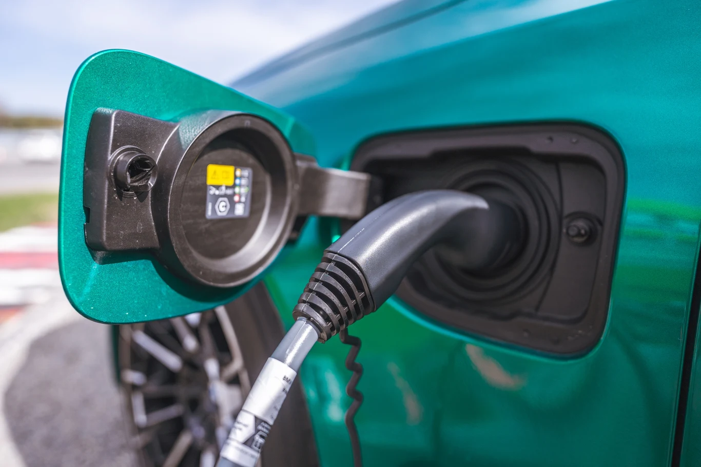 Wtyczka ładowania podłączona do portu elektrycznego samochodu, zbliżenie na szczegóły techniczne i lakier karoserii w kolorze turkusowym.