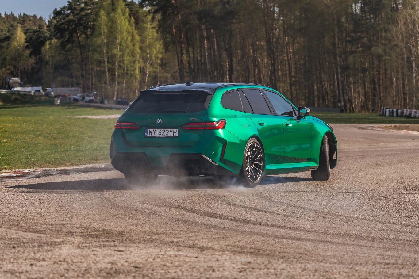 Zielony samochód marki BMW w trakcie dynamicznego skrętu na torze, widoczne unoszące się kłęby kurzu spod tylnej osi, otoczenie stanowi zieleń oraz las w tle.