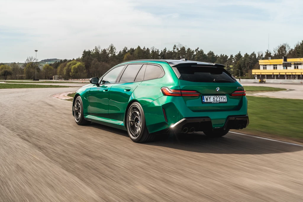 BMW M5 Touring obecnie niemal nie ma konkurencji. Ale czy to uzasadnia bazową cenę wynoszącą niemal 700 tys. zł? Zielone kombi BMW na torze wyścigowym, samochód dynamicznie pokonuje zakręt na asfalcie, w tle widoczny pas drzew oraz żółty budynek przy torze.