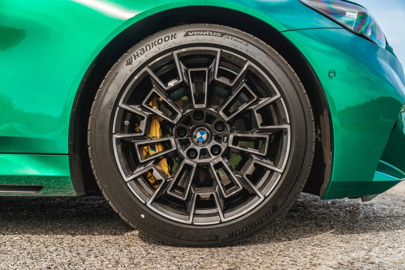 Felga aluminiowa o sportowym wzorze z logotypem BMW i żółtym zaciskiem hamulcowym, opona marki Hankook Ventus, karoseria samochodu w zielonym metalicznym kolorze.