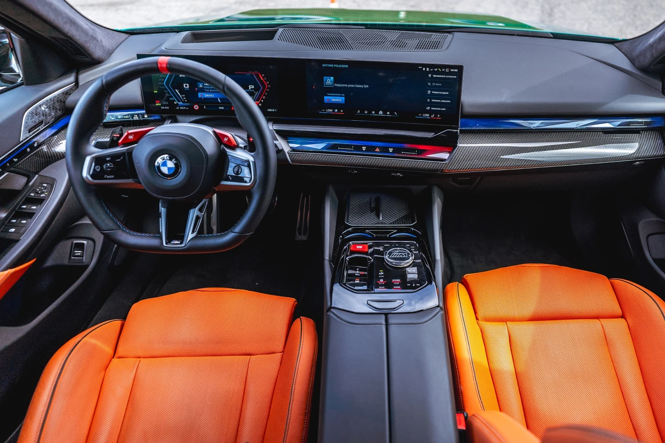 Luksusowe wnętrze samochodu BMW z nowoczesną deską rozdzielczą, ekranem multimedialnym, sportową kierownicą i skórzanymi pomarańczowymi fotelami.