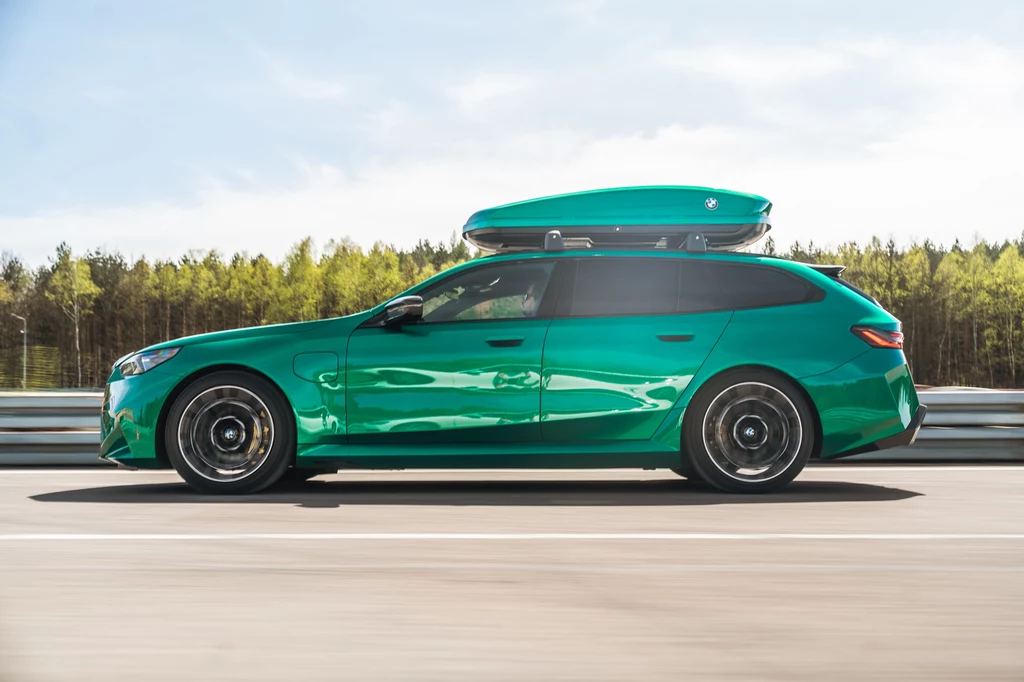 BMW M5 Touring ma prawie 5,1 m długości i prezentuje się bardzo masywnie Zielony samochód osobowy typu kombi w ruchu, z boxem dachowym, jedzie po drodze na tle lasu i niebieskiego nieba.