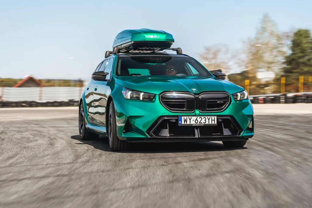 Wiele osób uważa, że nowa seria 5 ma dość kontrowersyjną stylistykę, ale M5 prezentuje się naprawdę dobrze Zielony samochód marki BMW z zamontowanym boksem dachowym porusza się po asfaltowej nawierzchni, w tle widoczne są rozmazane drzewa i ogrodzenie toru.