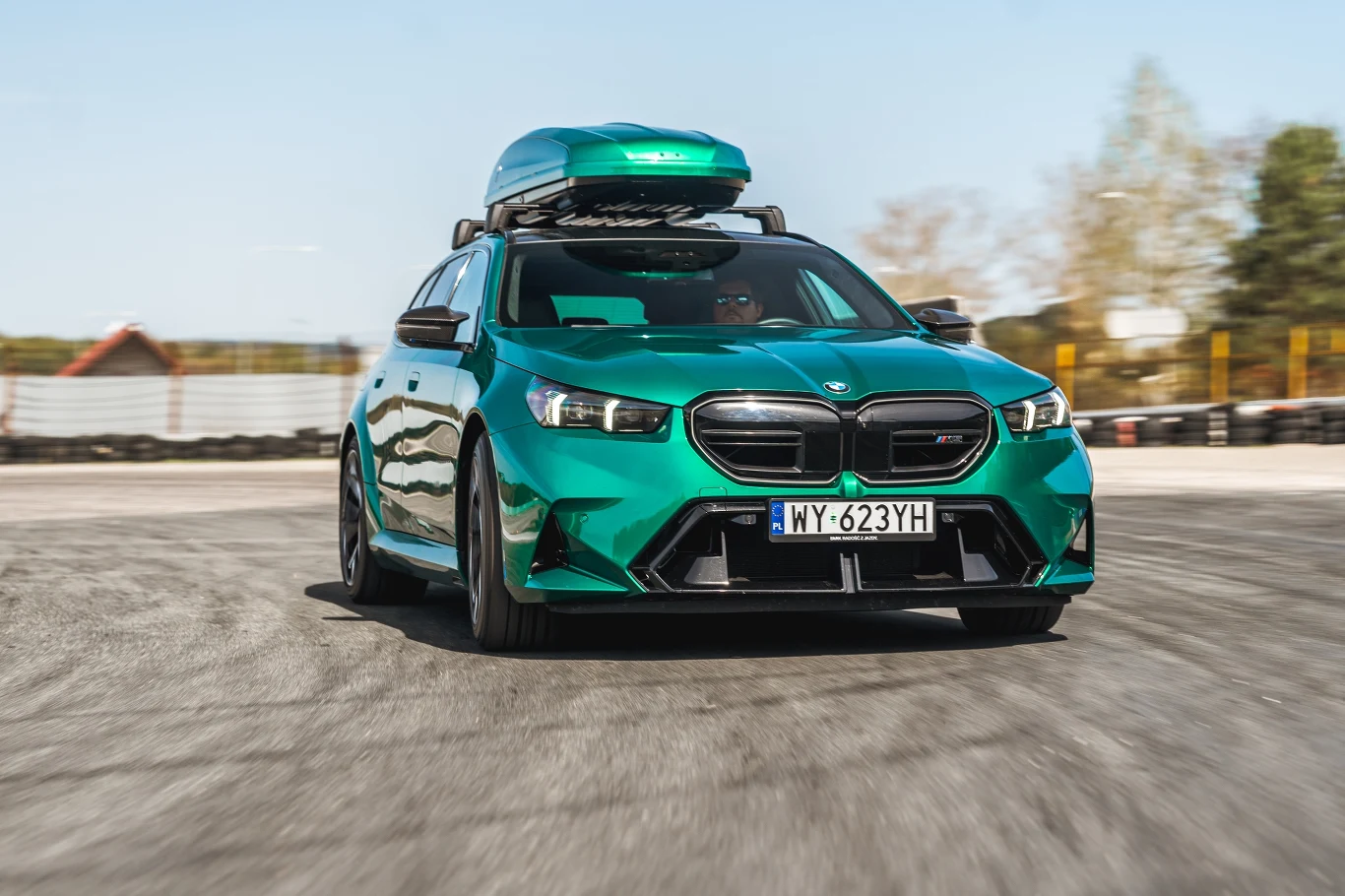 Zielony samochód marki BMW z zamontowanym boksem dachowym porusza się po asfaltowej nawierzchni, w tle widoczne są rozmazane drzewa i ogrodzenie toru.