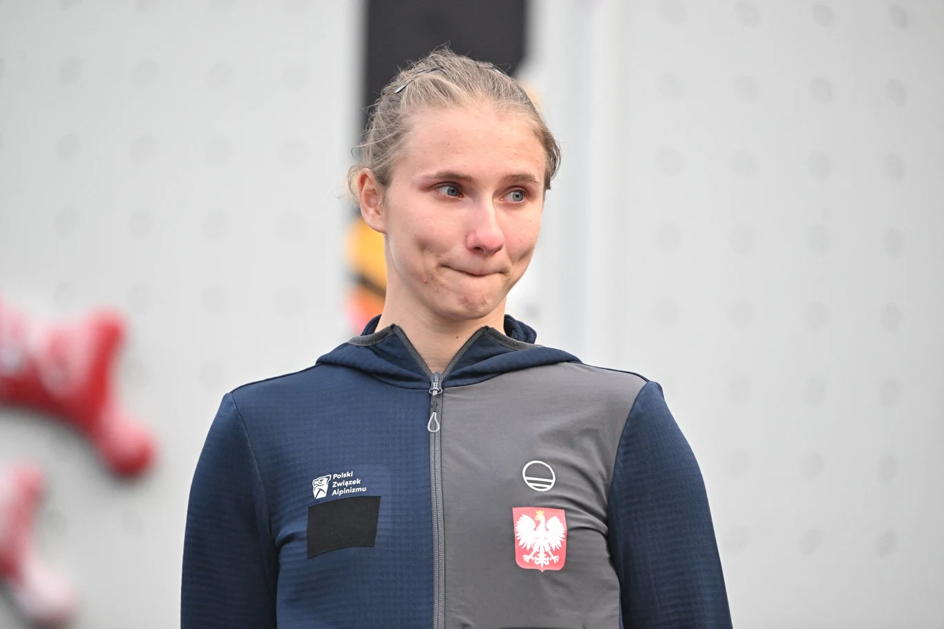 Natalia Kałucka Młoda kobieta w sportowej bluzie z polskimi emblematami stoi na tle ściany wspinaczkowej, wyraża skupienie lub emocje, jej włosy spięte są w kok.