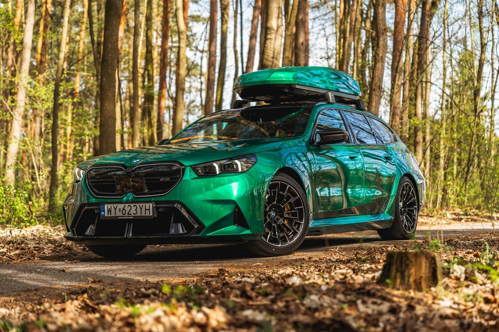 BMW M5 Touring Jasnozielony samochód terenowy z czarnymi detalami oraz pojemnym boksem dachowym stoi zaparkowany na leśnej drodze, otoczony drzewami i liśćmi na ziemi, podkreślając atmosferę podróży i przygody.