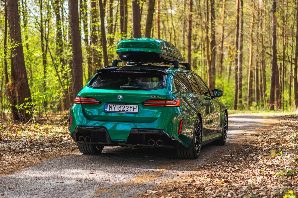 BMW M5 Touring Zielone BMW serii M z boxem dachowym parkuje na wąskiej asfaltowej drodze pośród wysokich drzew w lesie, otoczone przez bujną zieleń i liście opadłe na ziemi.