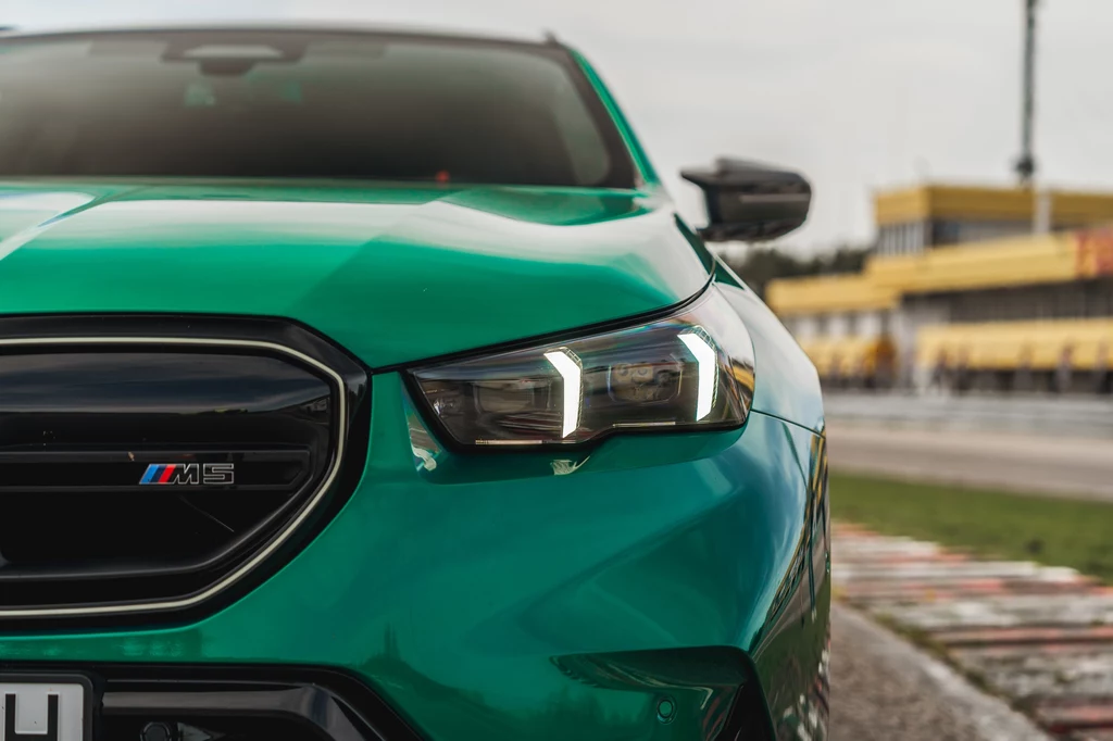 BMW M5 Touring Zbliżenie na przednią część zielonego samochodu sportowego marki BMW M8, z widocznym reflektorem LED i logo M8 na grillu, w tle rozmyta infrastruktura toru wyścigowego.