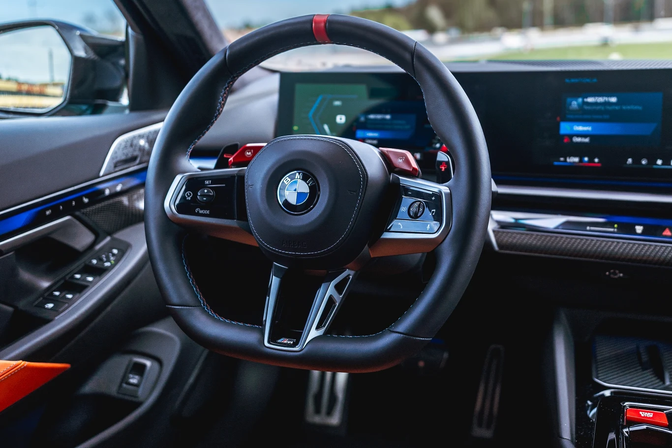 Nowoczesna kierownica z logo BMW, wyświetlacze cyfrowe oraz fragment konsoli centralnej z elementami sterowania, wszystko w sportowym, luksusowym wnętrzu samochodu.