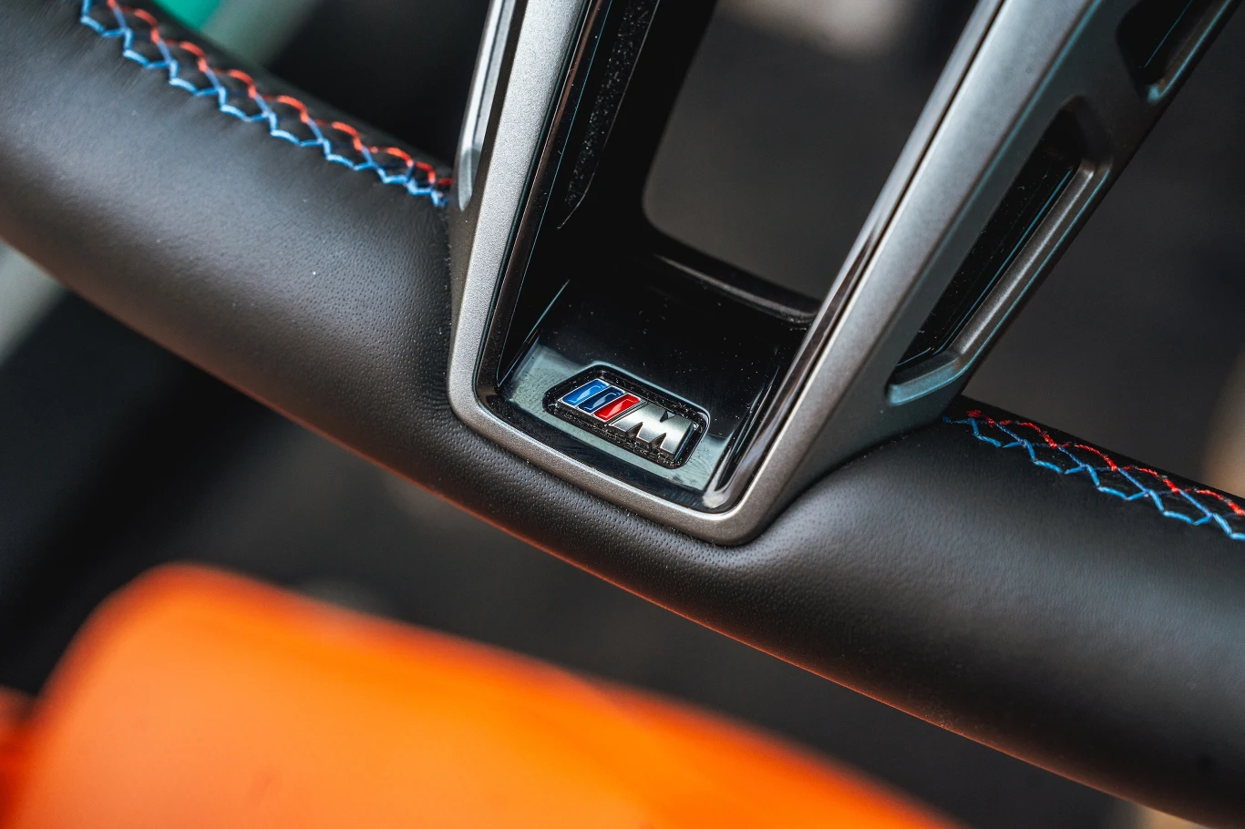 Czarny skórzany sportowy kierownica z kolorowymi przeszyciami oraz logo BMW M umieszczonym centralnie w dolnej części koła, widoczny fragment pomarańczowego fotela samochodowego.