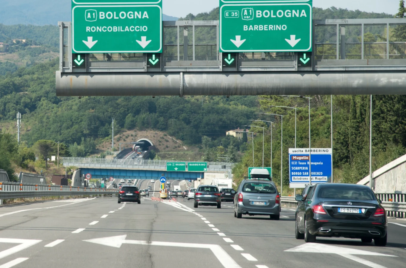 Jak wysokie są mandaty we Włoszech? Ruchliwa autostrada otoczona zielonymi wzgórzami, widoczne znaki drogowe kierujące do Bolonii przez Roncobilaccio i Barberino, liczne pojazdy poruszające się w obu kierunkach, dalszy plan z tunelem i lasem.
