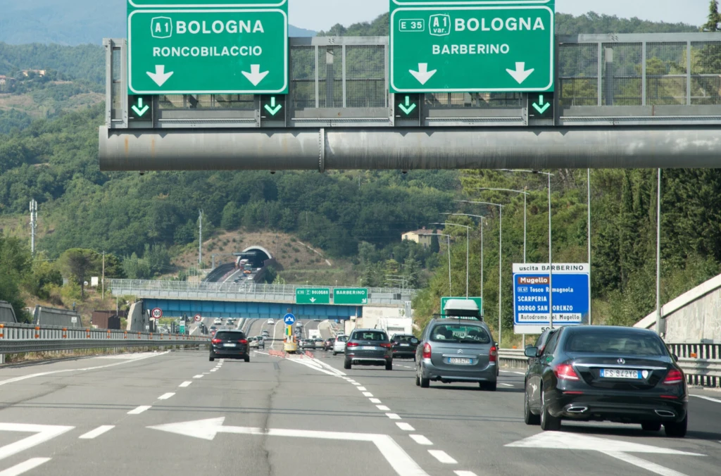 Ruchliwa autostrada otoczona zielonymi wzgórzami, widoczne znaki drogowe kierujące do Bolonii przez Roncobilaccio i Barberino, liczne pojazdy poruszające się w obu kierunkach, dalszy plan z tunelem i lasem.