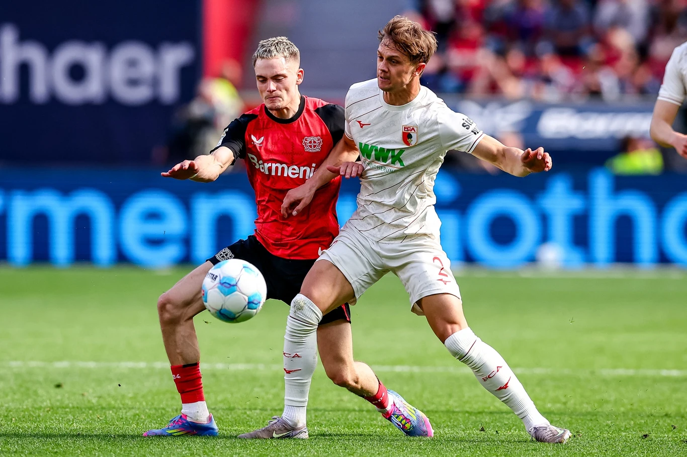 Robert Gumny w pojedynku z Florianem Wirtzem Dwóch piłkarzy walczy o piłkę podczas meczu na stadionie, jeden ubrany w czerwono-czarny strój, drugi w biały, obaj są zaangażowani w dynamiczne starcie o posiadanie futbolówki.