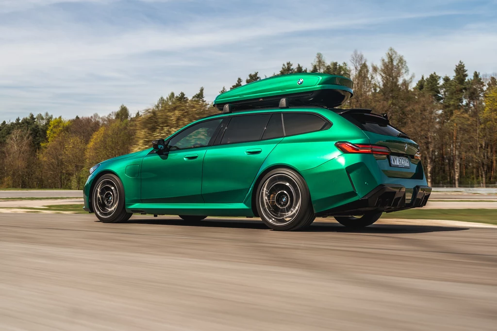 BMW M5 Touring świetnie prezentuje się w kolorze Zielony Isle of Man M. Co ciekawe, jest to lakier oferowany bezpłatnie zielone kombi z boxem dachowym jadące po drodze, dynamiczne ujęcie z tyłu, w tle drzewa i niebo z lekkim zachmurzeniem