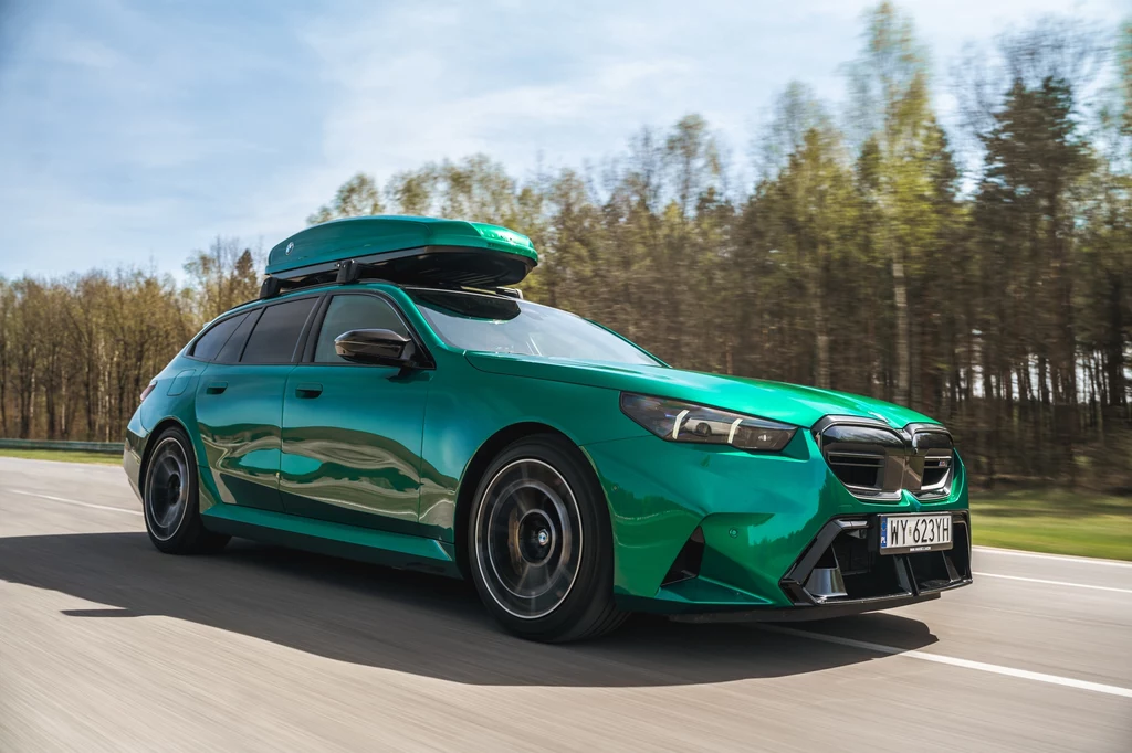 BMW M5 Touring Zielony samochód osobowy kombi z belkami dachowymi i boksem zamontowanym na dachu porusza się po drodze, w tle rząd drzew oraz niebieskie niebo z lekkim zachmurzeniem.