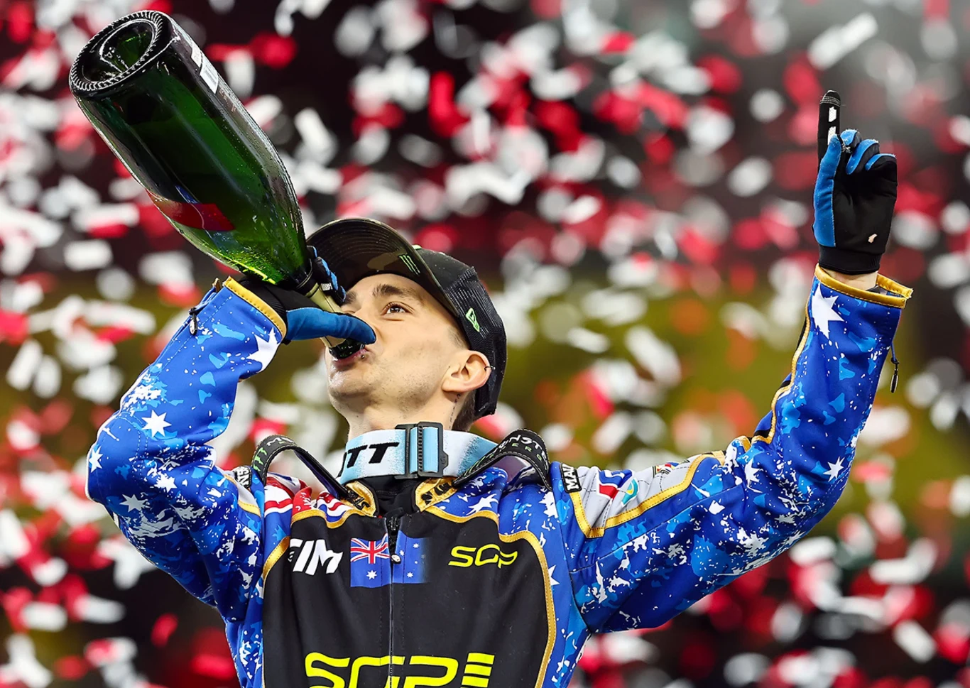 Zwycięzca wyścigu motorsportowego w stroju z motywem flagi Australii pije szampana z dużej butelki, w tle unoszą się czerwono-białe konfetti, tworząc uroczystą atmosferę.