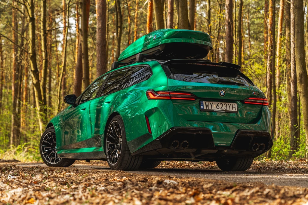 BMW M5 Touring Zielony samochód BMW kombi z czarnym bagażnikiem dachowym stoi zaparkowany na leśnej drodze, otoczony drzewami, światło słoneczne przebija się przez gałęzie