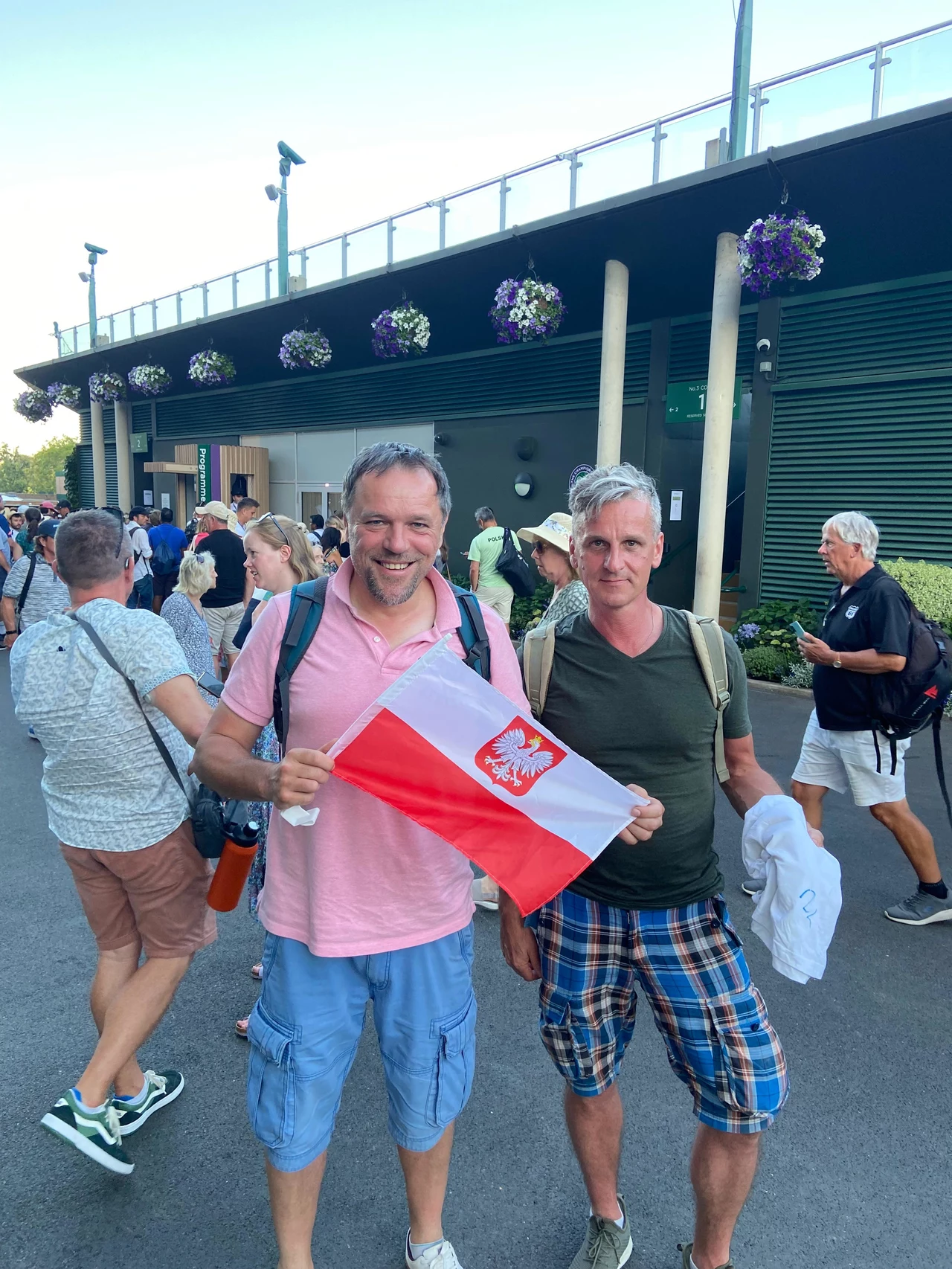 Remigiusz z Londynu (z lewej) i Tomek ze Świdnicy Dwóch mężczyzn w letnich ubraniach trzyma polską flagę, uśmiechając się do aparatu. W tle przechodzi grupa ludzi pod nowoczesnym budynkiem, nad którym wiszą kwiaty w doniczkach, panuje pogodna atmosfera.