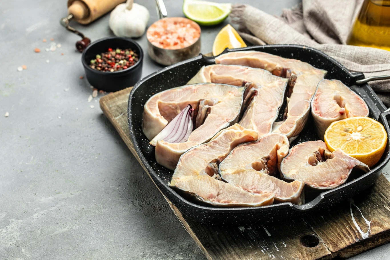 Surowe dzwonka ryby ułożone na patelni grillowej, obok połówka cytryny, zioła, przyprawy i oliwa widoczne na desce i blacie, akcesoria kuchenne w tle.