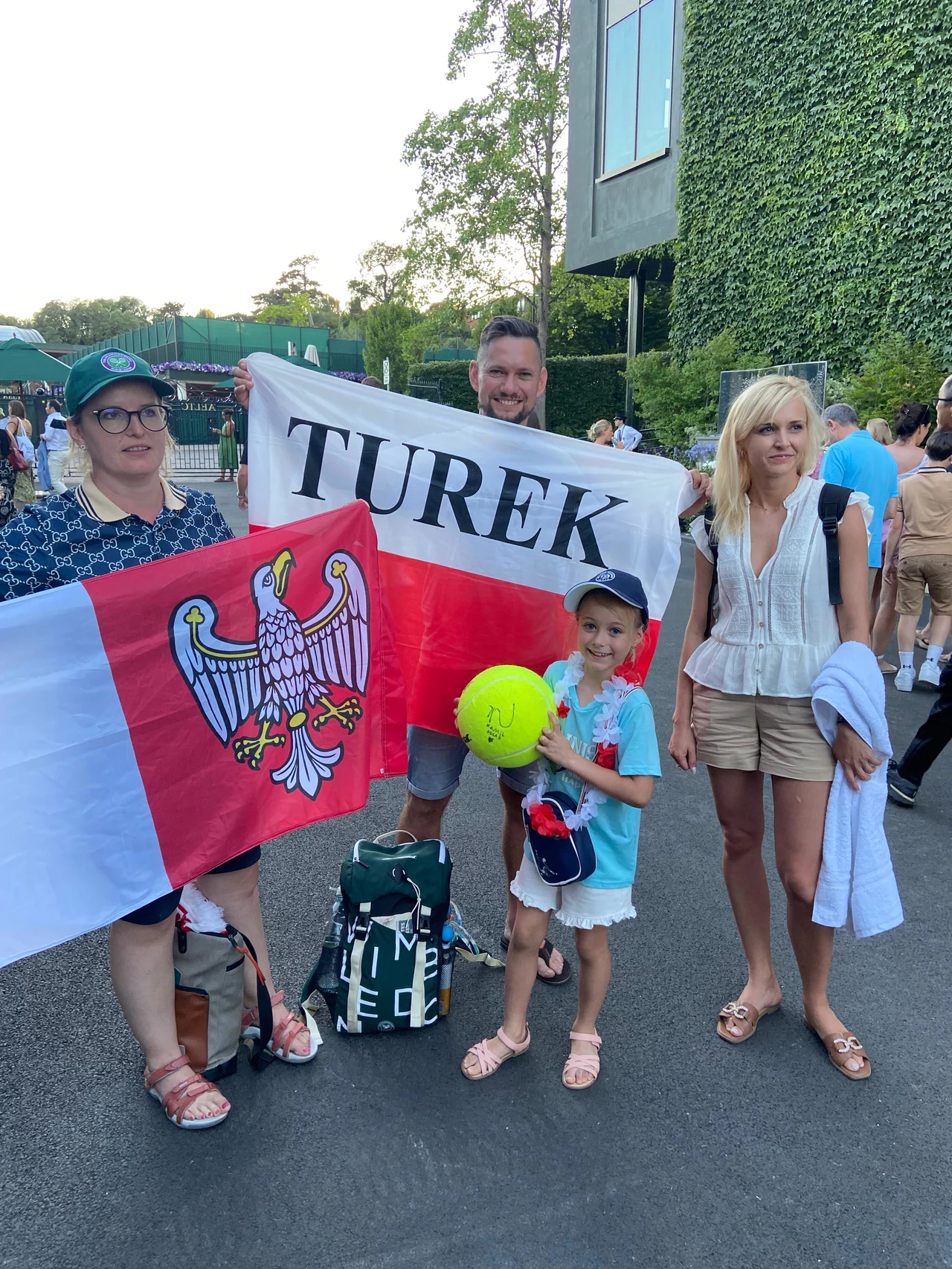 Od lewej: Anna, Dariusz, Monika i najmłodsza Lena. Na zdjęciu brakuje tylko Mateusza Grupa czterech osób, w tym dziecko trzymające dużą piłkę tenisową, pozuje z biało-czerwoną flagą z napisem Turek oraz flagą z orłem. W tle widoczne są osoby oraz budynek porośnięty bluszczem.