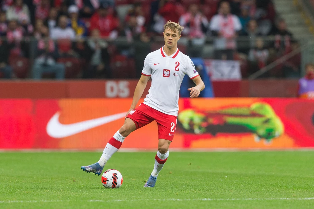 Robert Gumny w reprezentacji Polski Piłkarz reprezentacji Polski w białej koszulce i czerwonych spodenkach prowadzi piłkę na murawie stadionu, w tle widoczne trybuny z kibicami oraz barwne reklamy.