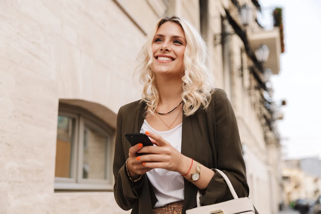 Koziorożec, Panna i Bliźnięta wiedzą, jak działać, gdy wszystko się wali Młoda kobieta z blond włosami uśmiecha się, trzymając w ręku smartfon, ubraną w elegancką marynarkę, białą bluzkę i jasną torebkę, stoi na tle miejskiej ulicy w jasnym świetle dziennym