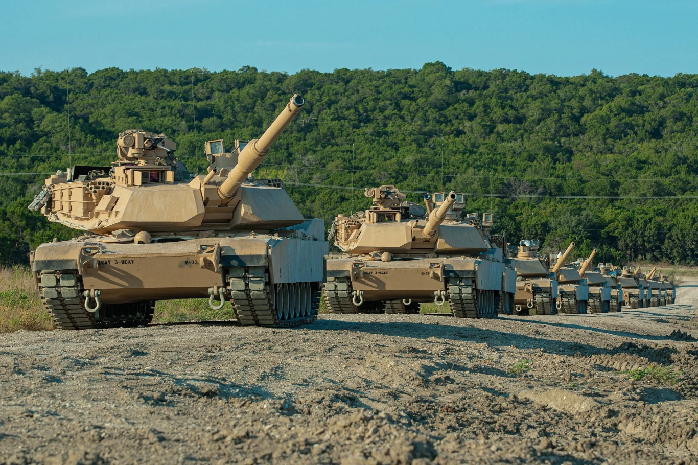 Czołgi M1A2 Abrams SEPv3 Kolumna czołgów ustawiona na drodze żwirowej na tle zielonego lasu pod niebieskim niebem, pojazdy znajdują się w równym szyku, każdy z lufą skierowaną do przodu.