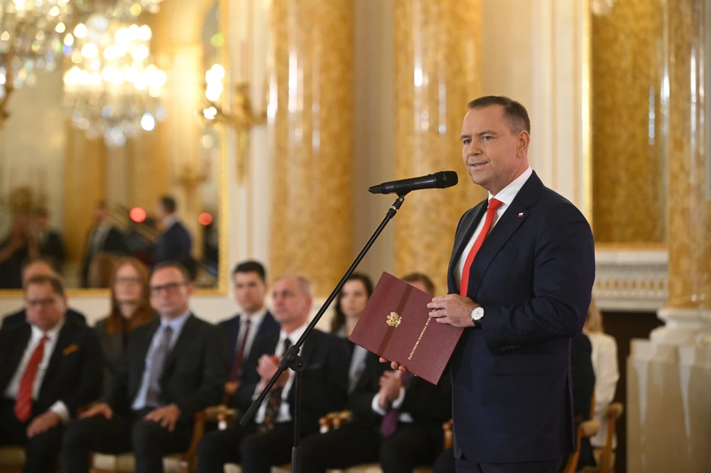 Karol Nawrocki podczas uroczystości wręczenia przez PKW zaświadczenia o wyborze na prezydenta Mężczyzna w garniturze z czerwoną krawatą stoi przy mikrofonie, trzymając w ręku dokument, w tle grupa osób siedzących w eleganckiej sali o złotych, zdobionych kolumnach.
