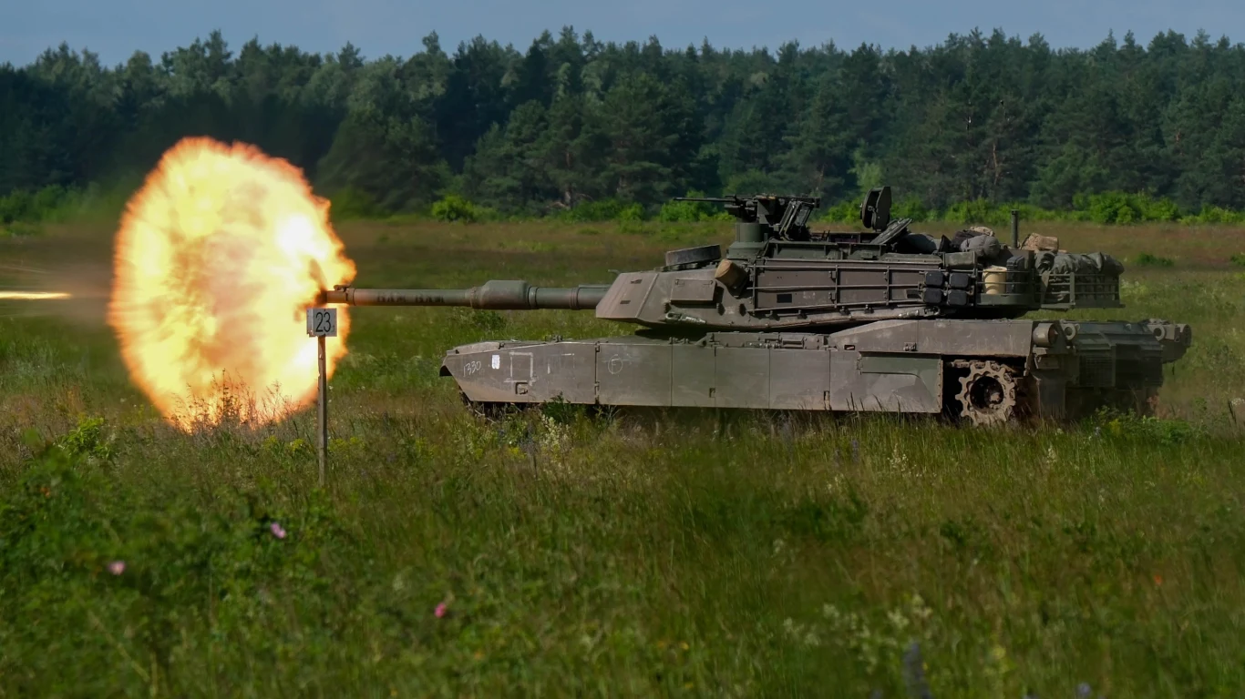 Amerykańskie Abramsy oddały strzały na poligonie Orzysz. Wielkie manewry sił NATO Czołg podczas oddawania strzału, eksplozja ognia widoczna przy jego lufie, tle zalesiony krajobraz i zielone pole.