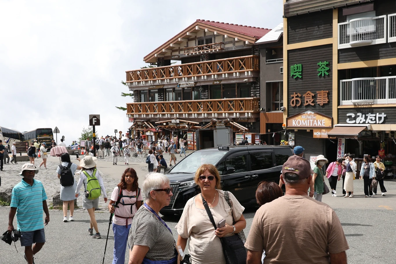 Tłum turystów przechadzających się po ulicy w górskiej miejscowości, okolica z nowoczesnymi drewnianymi budynkami i restauracjami z japońskimi napisami, z przodu zaparkowany czarny samochód.