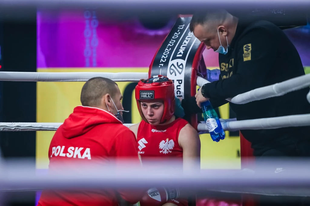 Bokserka w czerwonym stroju z napisem Polska siedzi w narożniku ringu podczas przerwy w walce, otoczona przez dwóch trenerów udzielających wskazówek i wsparcia.