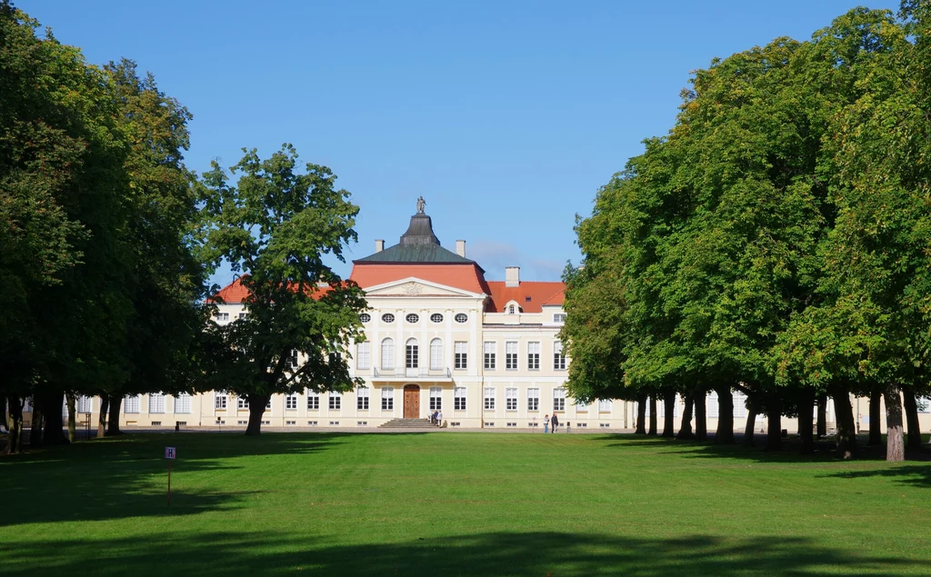 Pałac w Rogalinie to jeden z najwybitniejszych przykładów architektury rezydencjonalnej w Polsce Biały pałac z czerwonym dachem położony w głębi zadbanego parku, otoczony symetrycznym szpalerem drzew i szeroką trawiastą aleją prowadzącą do głównego wejścia.
