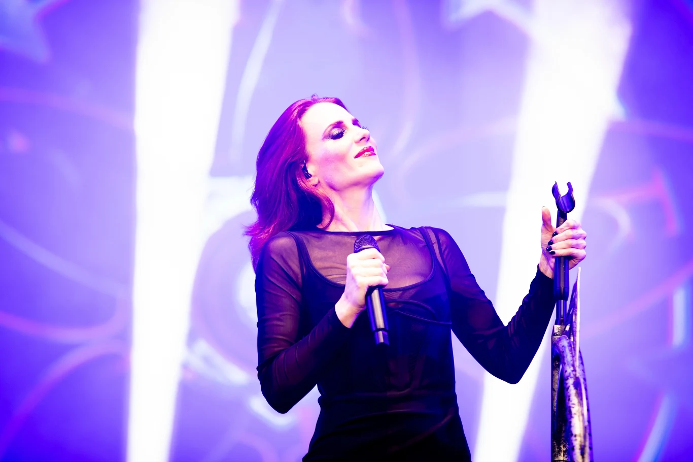Simone Simons (Epica) w akcji Kobieta w czarnej, półprzezroczystej bluzce śpiewa na scenie, trzyma mikrofon i statyw, otoczona jasnym, kolorowym światłem koncertowym.