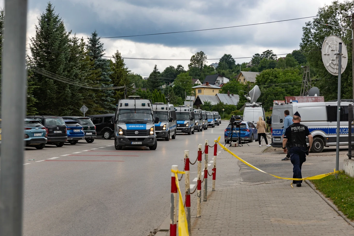 Obława w Starej Wsi Szpaler policyjnych radiowozów i wozów zabezpieczenia ustawionych wzdłuż ulicy, widoczne żółte taśmy odgradzające teren oraz funkcjonariusze pilnujący bezpieczeństwa w spokojnej, podmiejskiej okolicy.