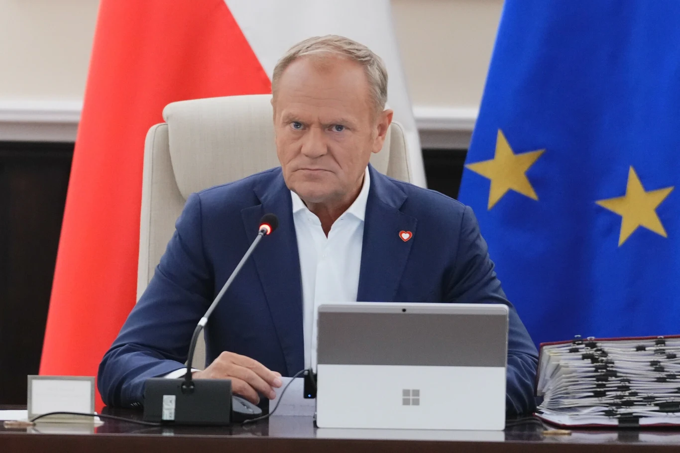 Premier Donald Tusk Mężczyzna w garniturze siedzi przy stole z mikrofonem, przed komputerem i dokumentami, w tle widoczne flagi Polski oraz Unii Europejskiej.
