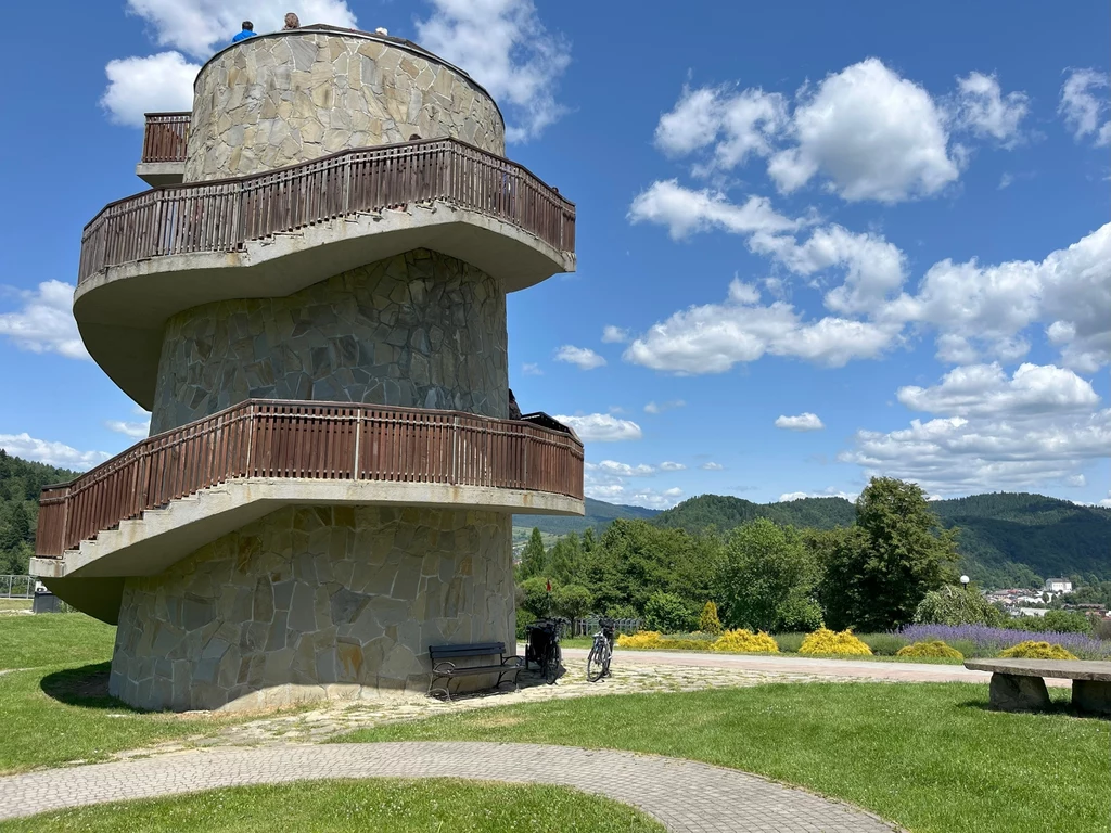 Wieża widokowa w Ogrodzie Sensorycznym. Roztacza się stąd panorama na całą okolicę Kamienna wieża widokowa ze spiralnymi schodami, otoczona zielonym trawnikiem i ławeczkami, w tle górski krajobraz z niebieskim niebem i białymi chmurami.