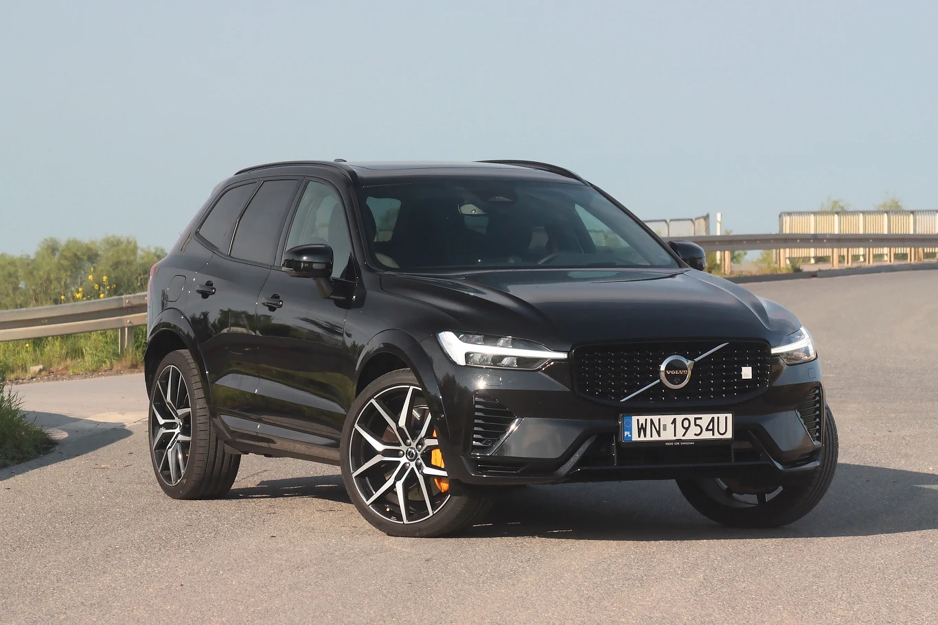 Czarny samochód Volvo SUV zaparkowany na asfalcie, widoczny z przodu pod lekkim kątem, z wyraźnym numerem rejestracyjnym na tle jasnego nieba oraz barierki ochronnej przy drodze.