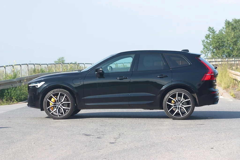 Volvo XC60 Polestar Enginereed Czarny SUV nowoczesnej marki stoi zaparkowany na asfalcie, w tle widoczny fragment barierki oraz drzewa, dominujące są kontrastujące ciemne felgi oraz żółte zaciski hamulców