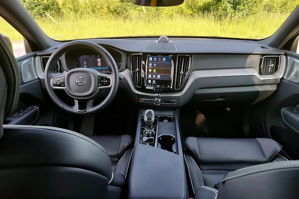 Volvo XC60 Wnętrze nowoczesnego samochodu marki Volvo z wyraźnie widoczną kierownicą, cyfrowym zestawem wskaźników oraz dużym ekranem dotykowym na centralnej konsoli, skórzane fotele i schludne, minimalistyczne wykończenie.