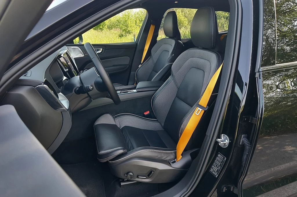 Volvo XC60 Wnętrze nowoczesnego samochodu z widocznymi przednimi fotelami pokrytymi czarną tapicerką ze sportowymi żółtymi pasami bezpieczeństwa, deska rozdzielcza z ekranem multimedialnym, szeroka szyba oraz drzwi kierowcy otwarte na zewnątrz.