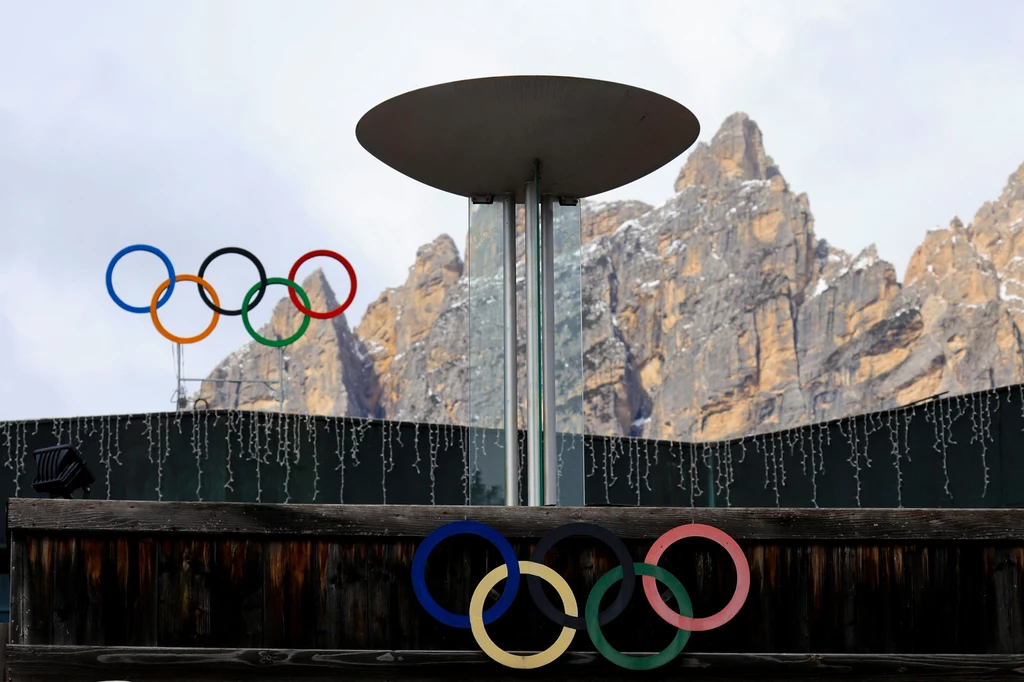 Cortina d'Ampezzo Stadion z olimpijskim zniczem i dwoma zestawami kolorowych kół olimpijskich na tle gór i ozdobnych lampek.