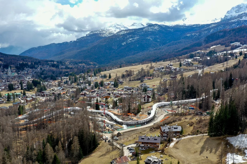 Budowa toru bobslejowo-saneczkowego w Cortinie d'Ampezzo Górska panorama z widokiem na miasteczko, zabudowania i obiekty sportowe pośród lasów i pól, w tle ośnieżone szczyty górskie oraz częściowo zachmurzone niebo.