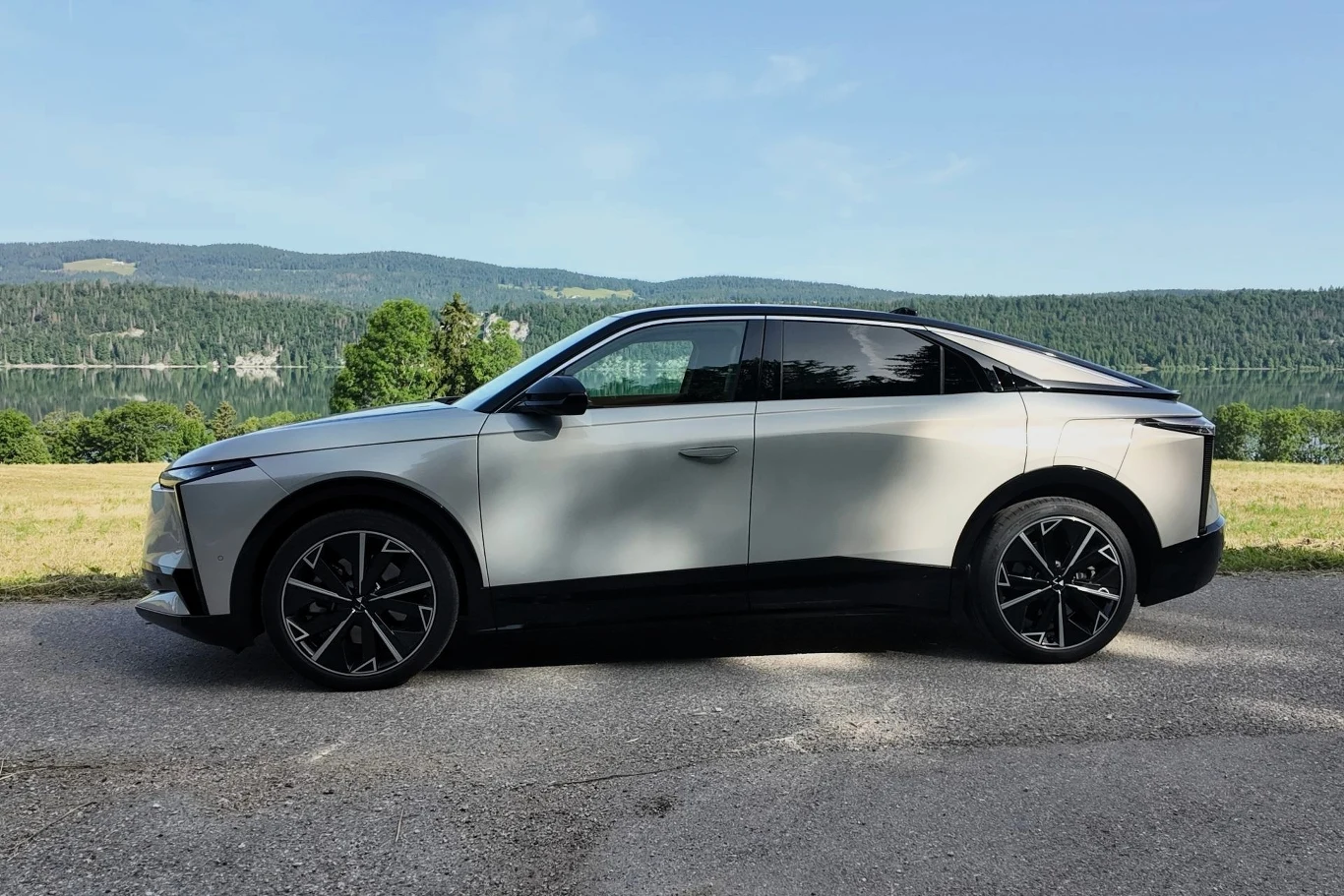 Nowoczesny, srebrny samochód osobowy o futurystycznej sylwetce zaparkowany na drodze, w tle zielony krajobraz z lasami i wzgórzami pod niebieskim niebem.