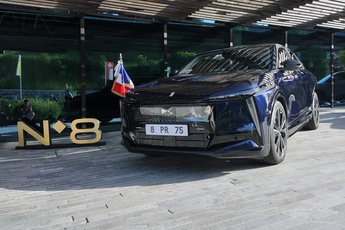 Nowoczesny, czarny samochód luksusowy zaparkowany na tle przeszklonego budynku, przyozdobiony małą francuską flagą, obok numer 8 wykonany z metalu jako dekoracja.