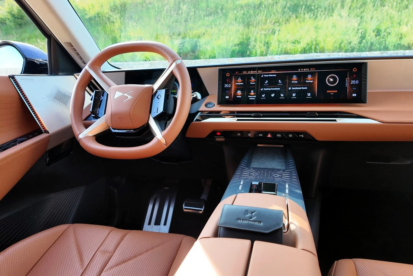 Wnętrze nowoczesnego samochodu z luksusową, brązową tapicerką skórzaną, dużym dotykowym ekranem multimedialnym oraz futurystyczną kierownicą, z widokiem na zielone otoczenie za szybą.