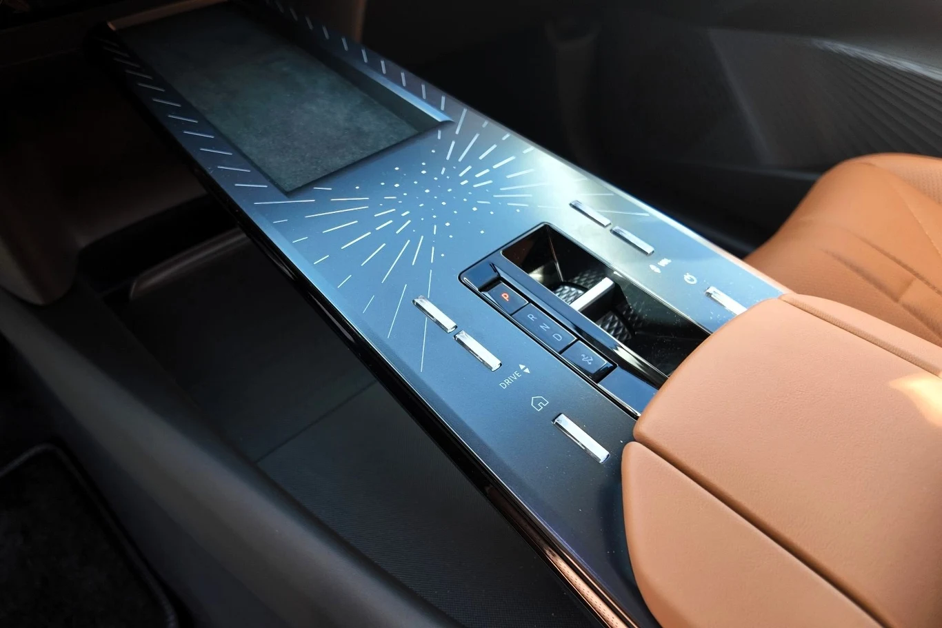 Nowoczesna konsola środkowa samochodu z czarno-srebrzystą powierzchnią, ozdobiona geometrycznymi wzorami, z przyciskami do sterowania funkcjami pojazdu oraz jasnym skórzanym podłokietnikiem.