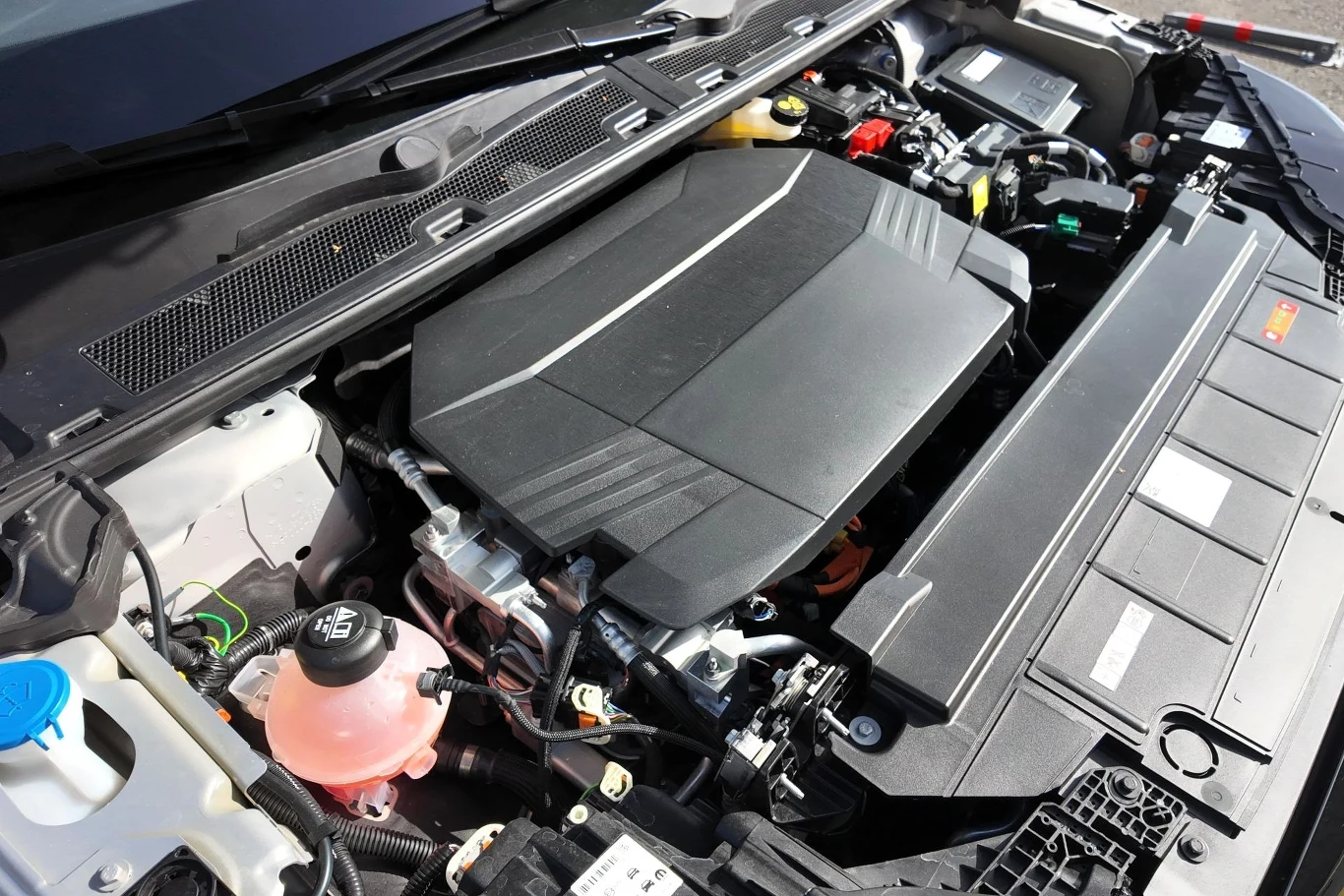 Silnik samochodowy z widocznymi elementami układu chłodzenia, zbiornikiem płynu oraz pokrywą silnika umieszczoną centralnie w komorze silnika.